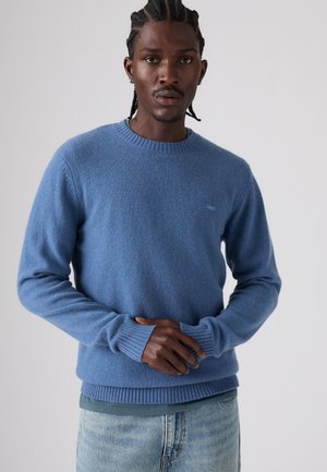 Homme aux cheveux tressés portant un pull bleu et un jean bleu clair, debout, les mains jointes devant un fond uni.