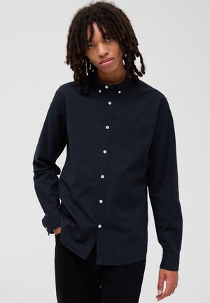 Chemise bleu marine à manches longues, avec col et boutons blancs. Texture lisse, coupe ajustée et détails de couture subtils sur les poignets.