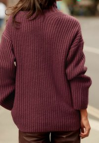 Grober Strickpullover in tiefem Burgunderrot mit hohem Kragen und lockeren Ärmeln, der ein strukturiertes Rippenmuster aufweist. Darunter sind braune Lederhosen sichtbar.