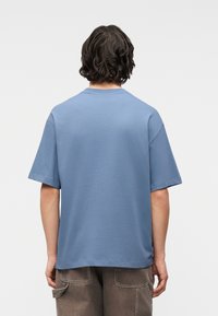 Άτομο με μεσαίου μήκους σκούρα μαλλιά που φοράει φαρδιά μπλε μπλούζα και καφέ παντελόνι με τσέπες, φαίνεται από πίσω σε απλό φόντο.