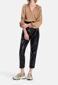 Femme portant un chemisier à manches longues beige, un pantalon en cuir noir taille haute et des baskets beiges, debout devant un fond blanc.