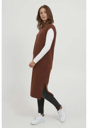 Braunes gestricktes ärmelloses Kleid mit seitlichen Schlitzen, getragen über einem weißen Langarmshirt, kombiniert mit schwarzen Jeans und weißen Sneakers.