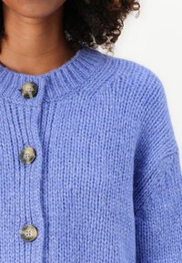 Šviesiai violetinė megzta kardiganas su grioveliu, apvali iškirpte ir trimis dekoratyvinėmis mygtukais priekyje.