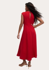 Vestido rojo con mangas en forma de capa, hasta la rodilla, con textura suave, detalle de ojal en la espalda, combinado con sandalias desnudas con tiras.