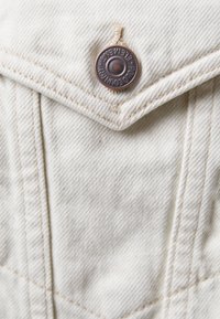 Nærbillede af beige denimstof med detaljeret syning og en kobberknap mærket "POLOCOUNTRY ELEMENT."