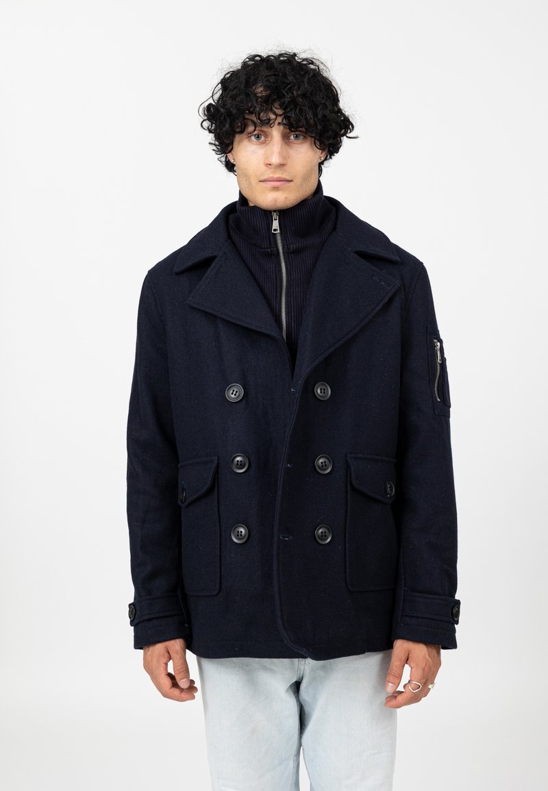 Granatowy dwurzędowy płaszcz typu peacoat z dużymi guzikami, dwiema przednimi kieszeniami i kieszenią na suwak na rękawie. Noszony na czarnym swetrze z wysokim kołnierzem.