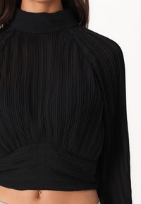 Blusa nera plissettata con collo alto e maniche lunghe. Il tessuto presenta un aspetto morbido e strutturato, con un dettaglio in vita arricciato.