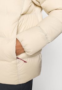 Beige Steppjacke mit gestepptem Design, ausgestattet mit einer Reißverschlusstasche mit rotem und blauem Kordelzug sowie einer glatten, weichen Textur.