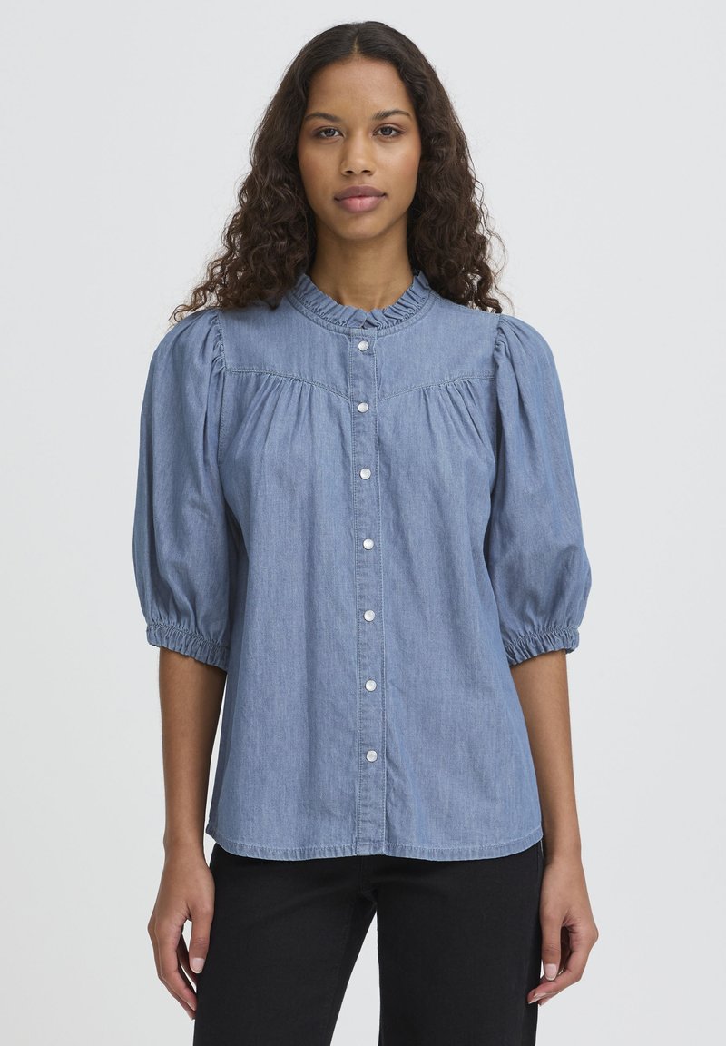 Lichtblauwe chambray blouse met opgeblazen mouwen, een gerafelde halslijn en witte knopen. De stof heeft een gladde textuur en een ontspannen pasvorm.