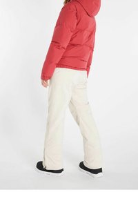 Veste matelassée rouge avec capuche, manches élastiques, associée à un pantalon beige clair avec fermetures éclair aux chevilles et chaussures noires.
