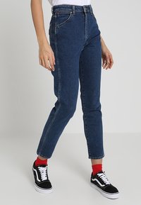Person som bär högmidjade mörkblå denimjeans, röda strumpor och svart-vita sneakers, som står framför en plain grå bakgrund.