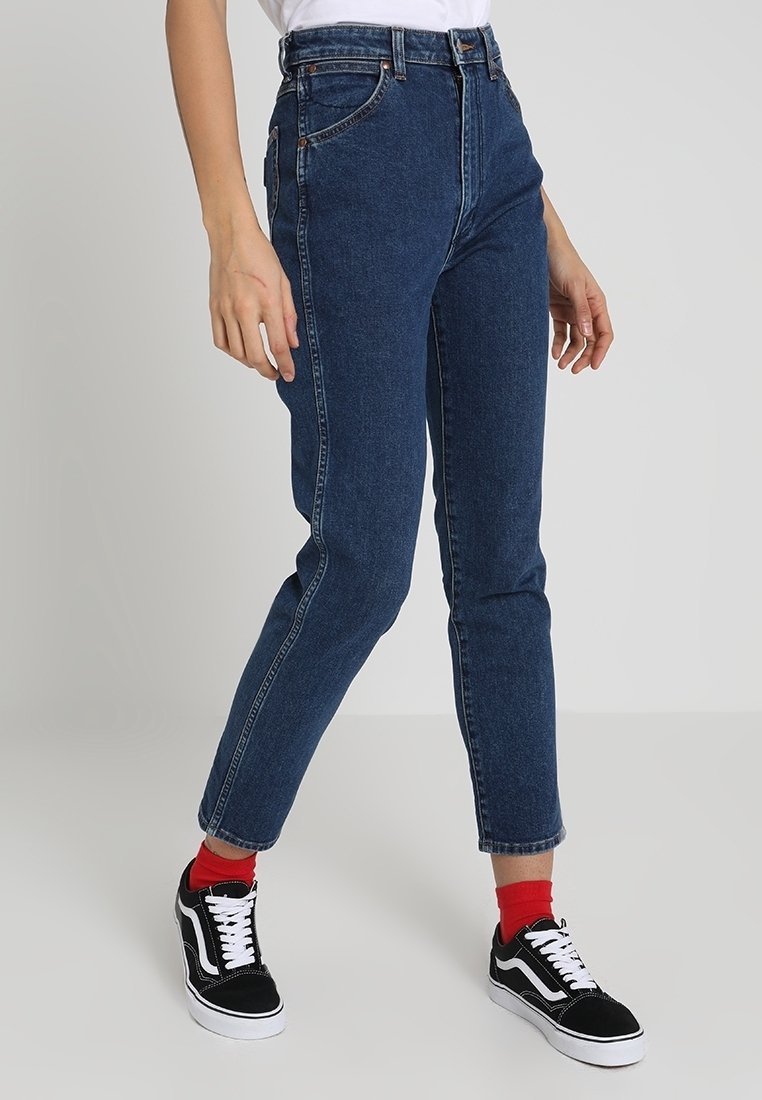 Person som bär högmidjade mörkblå denimjeans, röda strumpor och svart-vita sneakers, som står framför en plain grå bakgrund.