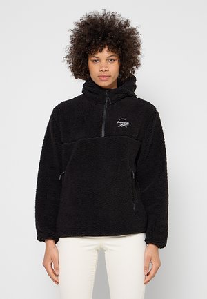 Person trägt schwarzen Reebok Fleece-Pullover mit halbem Reißverschluss und hohem Kragen, kombiniert mit weißen Hosen, steht vor einfarbigem Hintergrund.