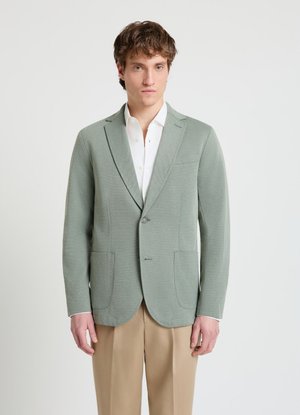 Mann trägt einen hellgrünen Blazer über einem weißen Hemd und beigen Hosen, steht vor einem einfarbigen hellen Hintergrund.