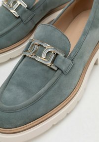 Grüne Veloursleder-Slipper mit weißen, klobigen Sohlen und goldener, dekorativer Metallschnalle oben.