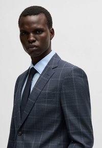 Completo a quadri blu navy con una vestibilità strutturata, abbinato a una camicia azzurra chiara e una cravatta scura; presenta un solo bottone e revers a punta.