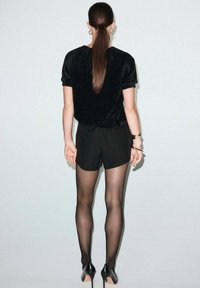 Top de manga corta de peluche sintético negro combinado con pantalones cortos negros de tiro alto y medias negras transparentes. Lleva zapatos negros de tacón bajo. Fondo simple.