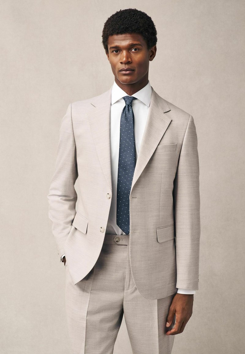 Traje de lino beige con solapas de muesca y dos botones, combinado con una camisa blanca y una corbata de lunares azul oscuro. Textura suave y corte entallado.