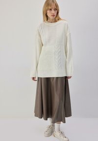 Pull en maille crème avec textures variées, associé à une longue jupe marron en similicuir. Le mannequin porte des chaussettes blanches et des baskets beiges.