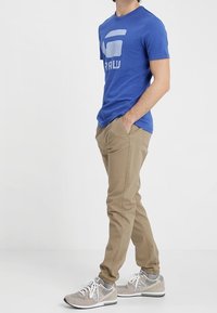 Mężczyzna w niebieskiej koszulce z grafiką, beżowych spodniach dresowych i szarych trampkach stoi z rękami w kieszeniach na tle białym.