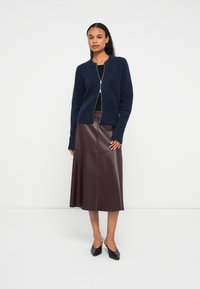 Moteris, vilkinti tamsiai mėlyną užtrauktuku užsegamą megztinį, juodą palaidinę, tamsiai rudą aukščiau kelių odinę sijoną ir juodas batų su žemu kulnu, stovi prieš balto fono.