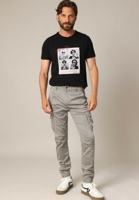 Man met baard, gekleed in een zwart graphic T-shirt met skeletfoto's, lichtgrijze cargobroek en witte sneakers, staande op een neutrale achtergrond.