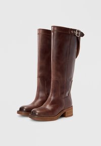 Καφέ δερμάτινες μπότες μέχρι το γόνατο με τετράγωνη μύτη, ξύλινο τακούνι σε σχήμα μπλοκ και πλάγιο πόμολο για ρύθμιση. Ομαλή υφή και διακριτική γυαλάδα.