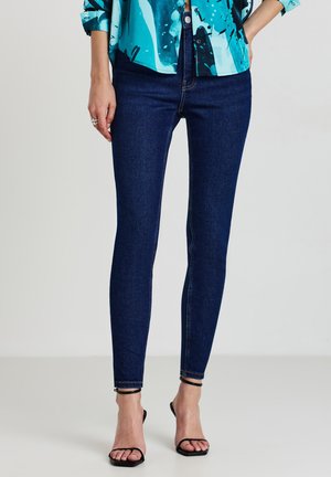Jeans ajustés de couleur bleu foncé en denim avec une texture lisse. Présentent une taille haute et des poignets à la cheville, associés à des sandales à talons noirs.