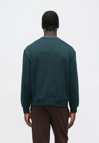 Sötét türkizkék pulóver kerek nyakkal, bordázott mandzsettákkal és szegéllyel. Simogató tapintású, lazább fazon, barna nadrág fölött viselve. Hátsó nézetben bemutatva.