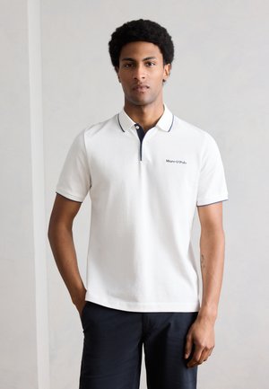 Férfi fehér, rövid ujjú, sötétkék szegélyű galléros pólóban és sötét nadrágban, egyszínű világosszürke háttér előtt állva.