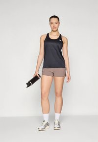 Camisola de atletismo preta sem mangas com logotipo, calções castanho claro, a segurar uma garrafa de água preta, usando meias brancas e sapatilhas cinzentas.