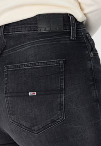 Vaqueros de mezclilla negra con una textura ligeramente desgastada. Presenta un bolsillo trasero rectangular y una etiqueta de cuero con el logo "TOMMY JEANS" grabado.