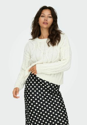 Pull en tricot blanc chunky avec un col rond et des poignets côtelés, associé à une jupe noire à pois, composée d'un tissu lisse.