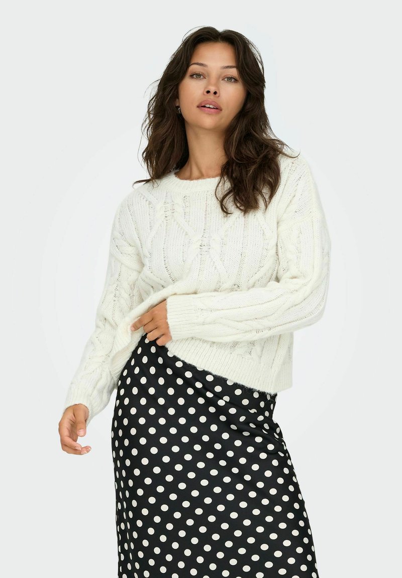 Fülliger weißer Kabelstrickpullover mit rundem Ausschnitt und gerippten Bündchen, kombiniert mit einem schwarzen Rock mit Punkten aus glattem Stoff.
