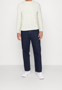 Cremefarbener, kabelgestrickter Pullover mit gerippten Bündchen und Saum, kombiniert mit dunklen, gerade geschnittenen Denim-Jeans und weißen Sportschuhen.