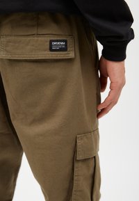 Olivgrüne Cargo-Hosen aus strapazierfähiger Baumwolle mit einer Gesäßtasche, die ein schwarzes DRDENIM-Label trägt. Sichtbare Handplatzierung auf dem Oberschenkel.