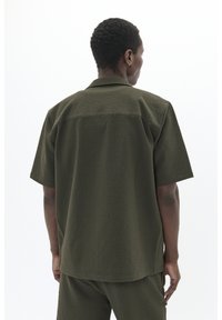 Camisa de botones de manga corta en tejido texturizado verde oscuro, con un cuello clásico y un ajuste relajado, vista desde la espalda.
