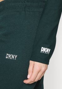 Tmavě zelený sportovní top s dlouhým rukávem, na kterém je stříbrné a bílé logo DKNY, žebrovaný výstřih a přiléhavé manžety. Měkká textilie.