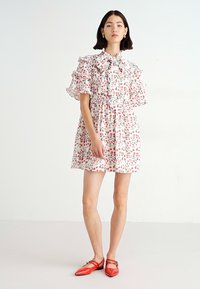 Vestido floral con mangas rizadas, un lazo en el cuello, en rosa con patrones de hojas verdes y rojas. Combinado con zapatos planos rojos, contra un fondo neutro.