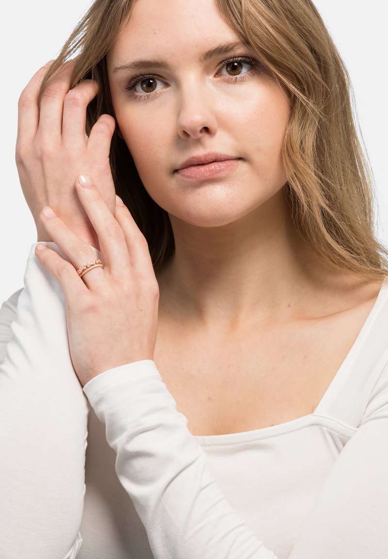 Gylden ring med delikat, sammenflettet mønster på en models finger, iført en tettsittende hvit topp med lange ermer mot en nøytral bakgrunn.