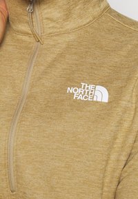 Fleece pulovr s polovičním zipem v světle hnědé barvě s kartáčovanou texturou. Má přední zip a bílé logo na levé části hrudníku.