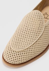 Mocassino in pelle intrecciata beige con punta arrotondata, dettaglio di cucitura e suola marrone chiaro su uno sfondo bianco.