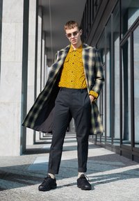 Kostkovaný kabát v černé a žluté barvě přes žlutou košili s leopardím vzorem, doplněný tmavými kalhotami a černými teniskami. Hladké textury, městské prostředí.