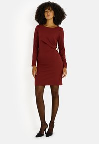 Robe moulante bordeaux à manches longues, décolleté arrondi, détail torsadé à la taille, associée à des chaussures à talons hauts noires.