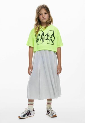 Fată tânără poartă un top scurt verde neon cu cifra „04,” o fustă plisată argintie strălucitoare, șosete dungi și adidași argintii, în fața unui fundal alb.