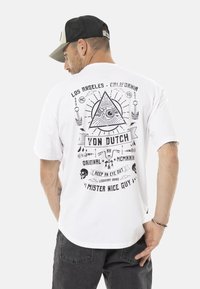 Camiseta de algodón blanca con estampado gráfico en negro que incluye un ojo, calaveras y texto. Cuello redondo, mangas cortas y ajuste relajado.