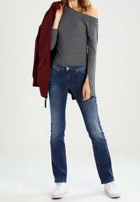 Top gris de manga larga y sin hombros combinado con vaqueros rectos azules y zapatillas blancas, con una chaqueta burdeos colgada de un hombro.