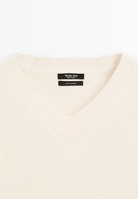 Maglione in cashmere a V nella tonalità beige chiaro. Presenta una texture a costine sul collo e sui polsini, con etichette che mostrano informazioni sul marchio e sul materiale.