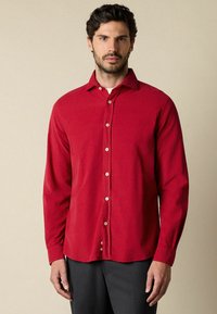 Man in een rood, knoop-gebonden, langemouwen shirt en donkergekleurde broek tegen een neutrale achtergrond, recht vooruit kijkend met een neutrale uitdrukking.
