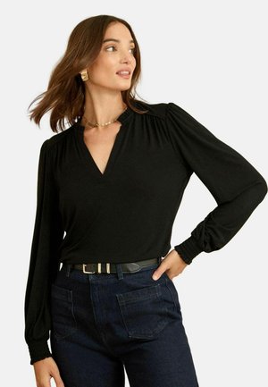 Schwarze Bluse mit langen Ärmeln und V-Ausschnitt, raffinierten Schulterdetails und figurbetonten Manschetten, kombiniert mit dunklen Jeans und einem schwarzen Gürtel.